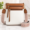 A structured crossbody bag with a white front body, tan leather top panel and trim, and a wide woven strap in white, tan, and rust tones. The bag has two zippered compartments on the top and is photographed on a light marble-like surface with a softly blurred indoor background.